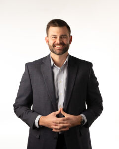 More casual headshot pose with hands clasped in front, man smiling at the camera, with beard on white background