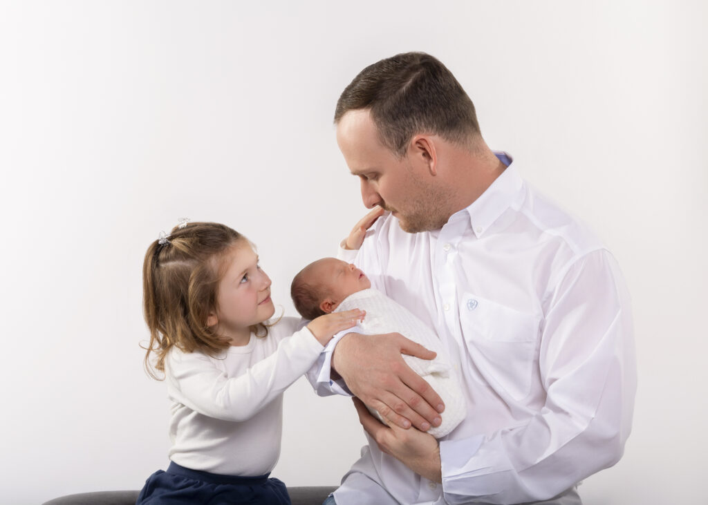 father's photoshoot Dad is holding his newborn daughter while his older daughter gazes at him lovingly. All wearing white. 