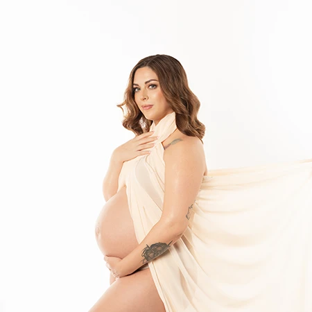 Fort Mill SC Mom to be, with dark wavy hair dressed in flowing fabric draped to cover her while showing off her exposed pregnant belly, glowing.