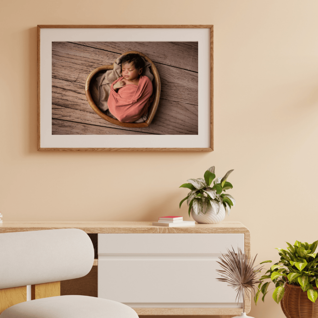  Over the desk framed newborn portrait of a dark skinned baby girl swaddled in coral peach fabric, lying in a wooden heart bowl. Portrait captured in Fort Mill Newborn Studio