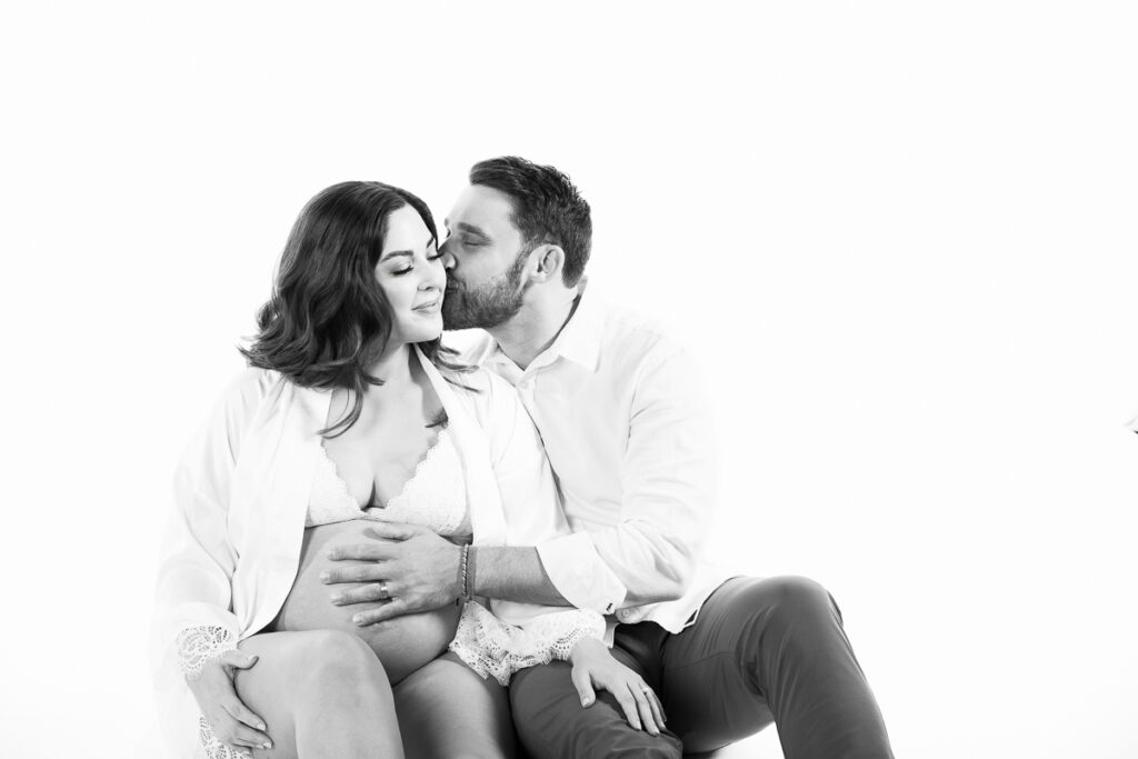 expecting Dad kissing mom to be on the cheek, no color, white backdrop allows the view to imagine this couple is relaxing at home preparing for a new baby.