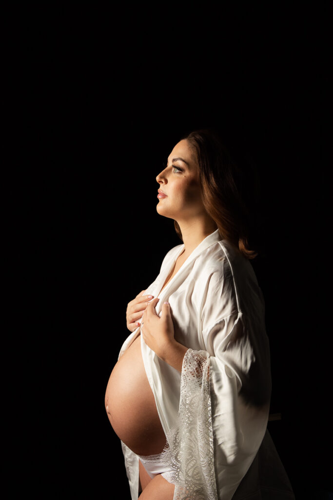 Expecting Charlotte, NC mom to be gazing away from the camera with a soft expression, showing her exposed pregnant belly wearing a white robe with lace trim. 