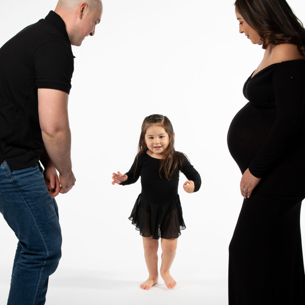 sweet toddler girl dancing between her pregnant mom and father all wearing black for a playful maternity portrait session Charlotte North Carolina