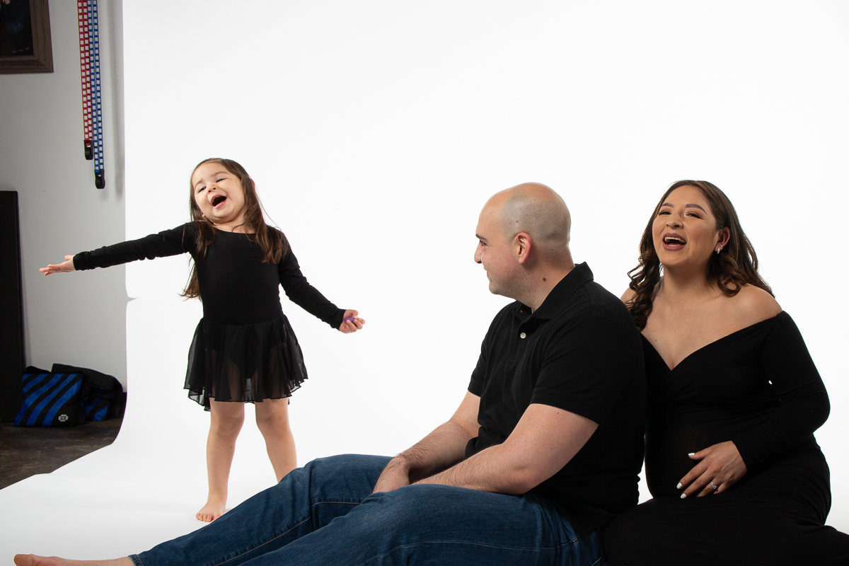 Silly toddler antics at this maternity pictures with toddlers portrait session, mom to be in laughing while dad to be looks at his daughter smiling