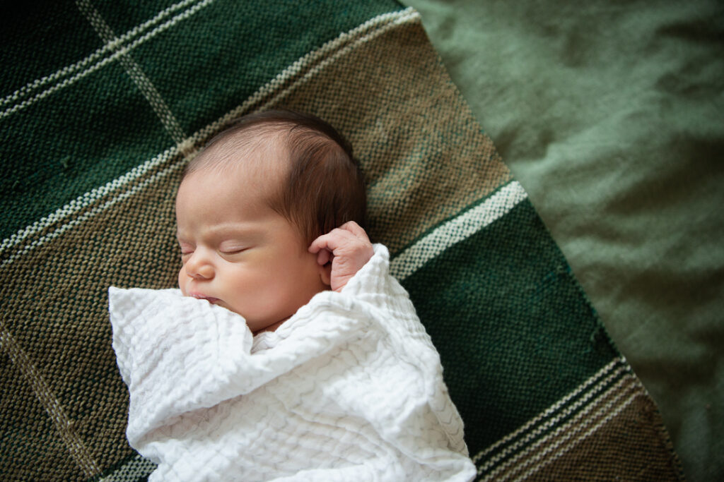 precious newborn sleeping cozily wrapped up on mom's bed for her In-Home Newborn Portraits in Fort Mill sc. baby is wrapped in white muslin blanket, laying on a green checked throw.