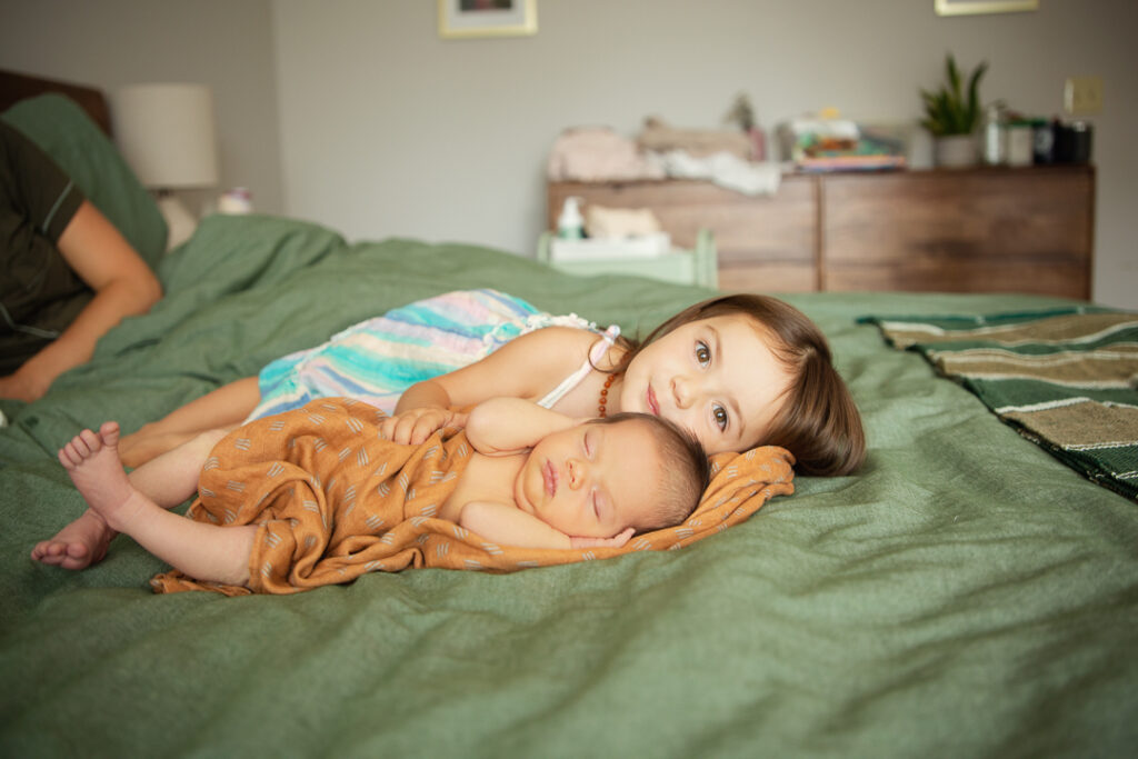 Precious baby girl wrapped in orange blanket, snuggled up with her big sister (2 years old) looking at the camera, both are in mom's bed and we see mom's arm in the corner. For these in-hone newborn portraits in Tega Cay SC