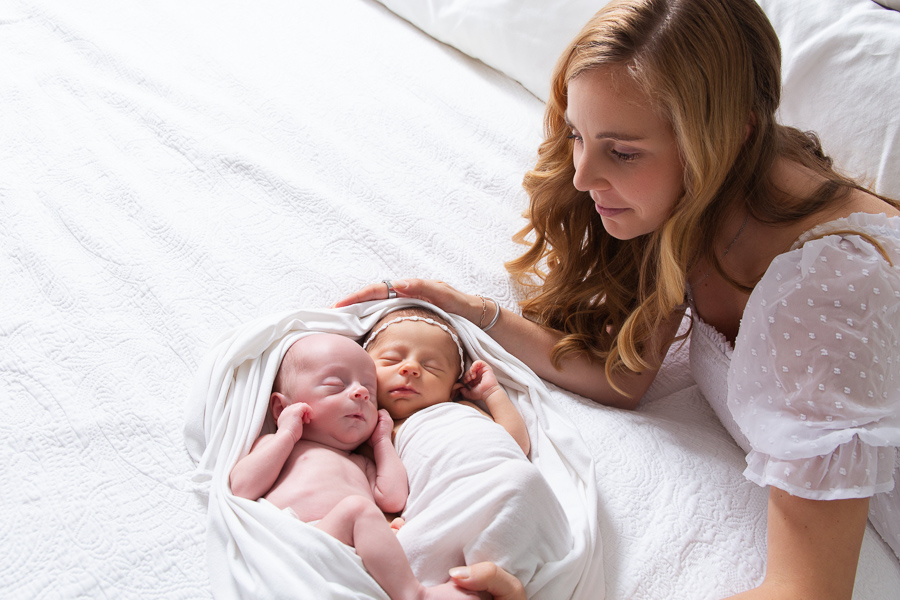 Charlotte newborn Portraits created in home Mom wearing a white dress is looking down at her twins baby girl and a baby boy gently swaddles in white, sleeping peacefully while she looks on.