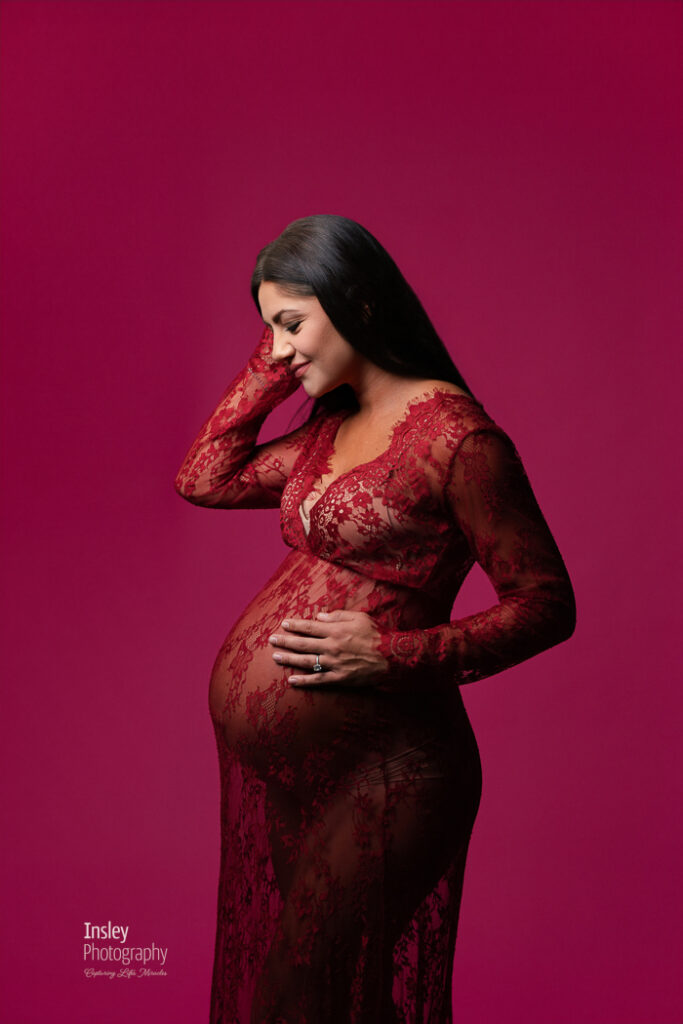 Pregnant woman in a deep red lace gown with her hand in her hair, looking down against a raspberry backdrop