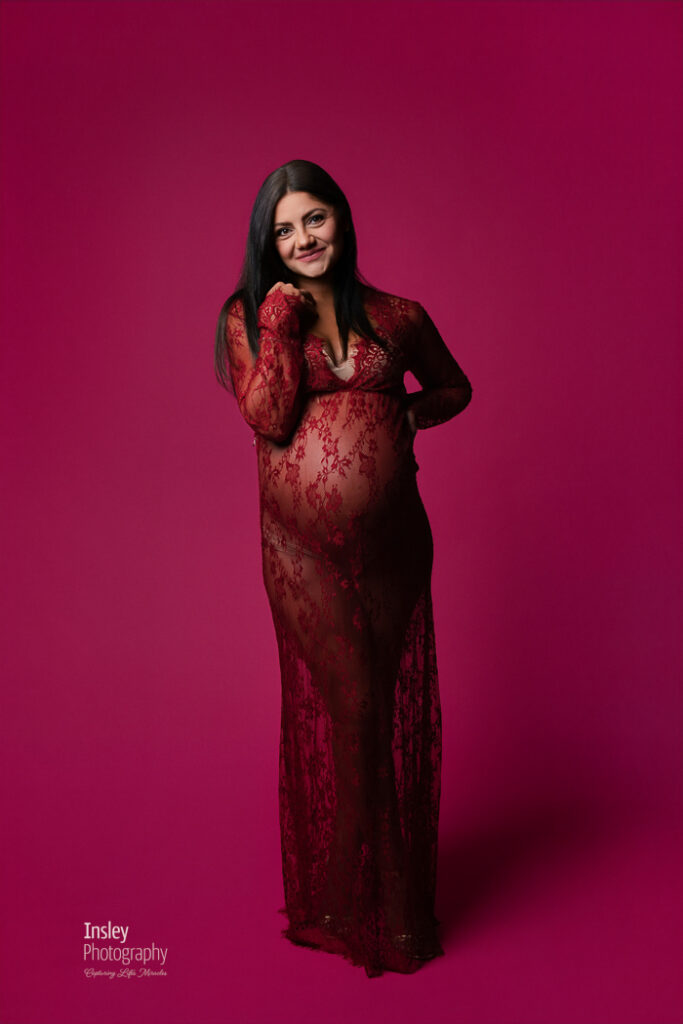 Full-length maternity portrait in a burgundy lace gown, expecting mother smiling on a deep pink backdrop, Fort Mill Studio session