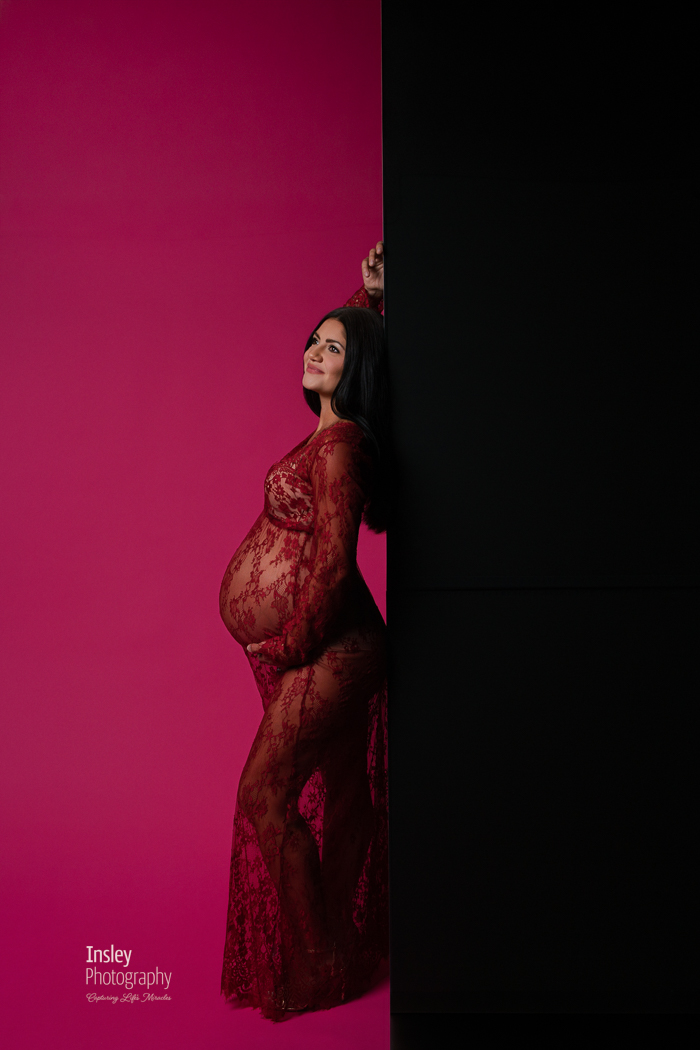 Expecting mother in red lace leaning against a black wall, dramatic maternity portrait with deep pink and black, created in the iNsley Photography studio in Fort Mill SC