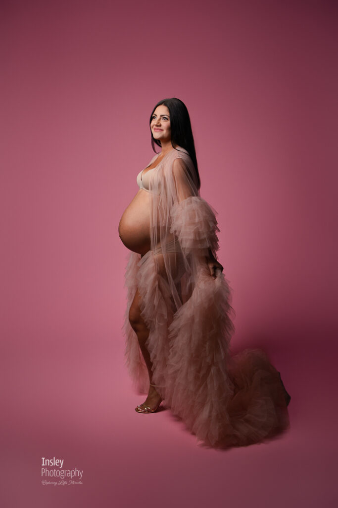 Pregnant woman in a blush tulle gown standing against a rose backdrop, full-length studio maternity portrait in Fort Mill SC