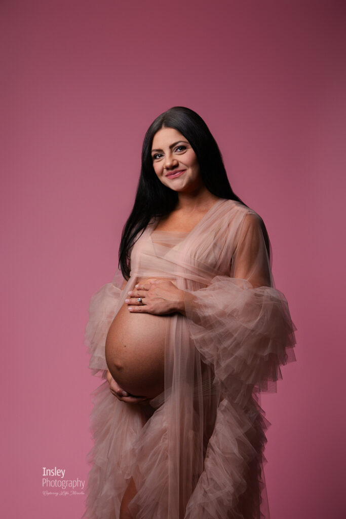 Expecting mother in blush tulle holding her belly and smiling at the camera, monochromatic pink maternity portrait