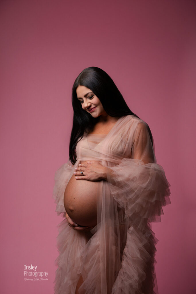 Pregnant woman looking down at her belly in a blush tulle gown, quiet moment from a pink studio maternity session