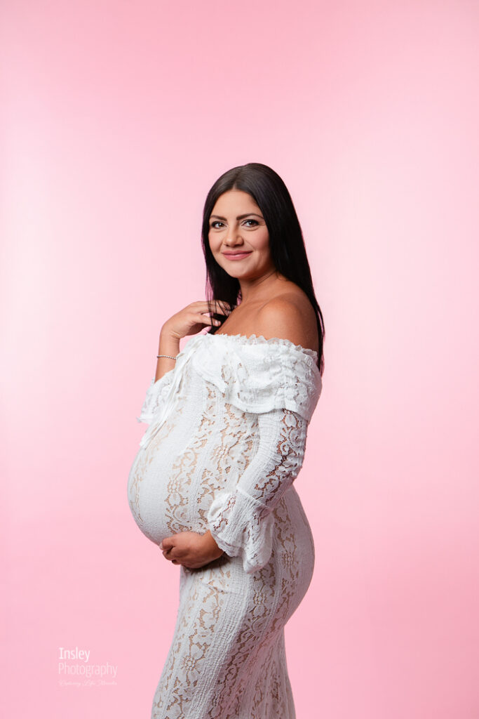 Expecting mother in a white lace off-shoulder gown against a soft pink backdrop, studio maternity portrait in Fort Mill SC