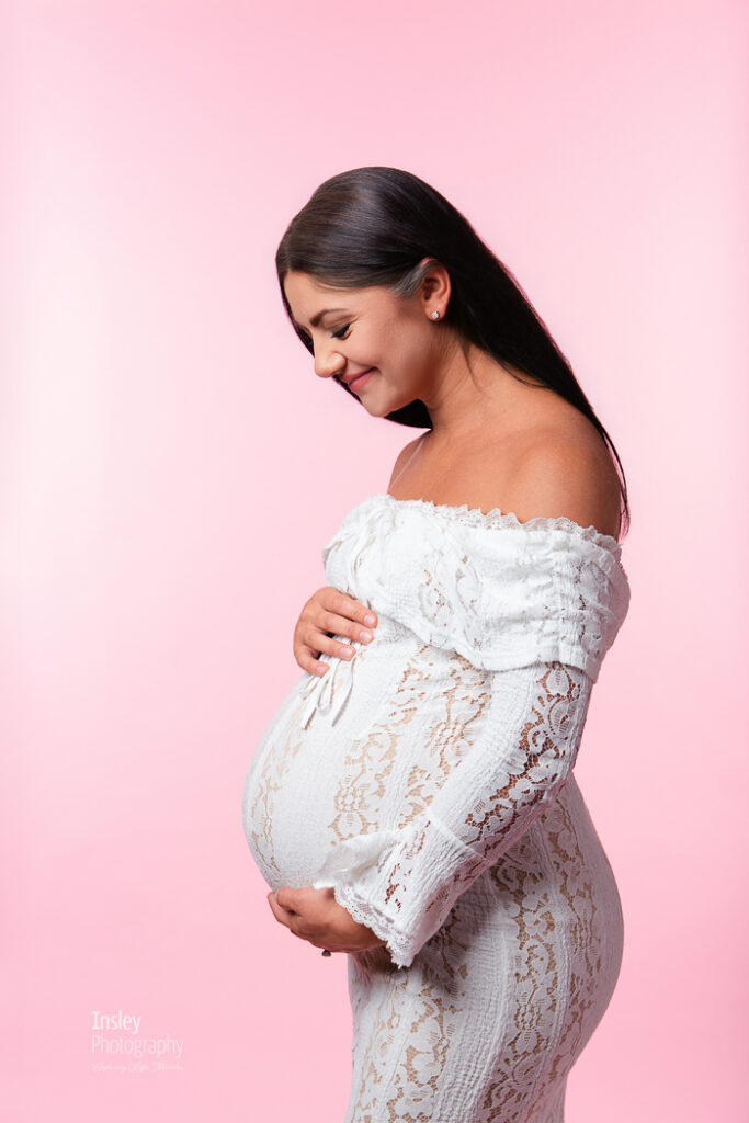 Pregnant woman looking down at her belly in a white lace gown, both hands cradling the bump