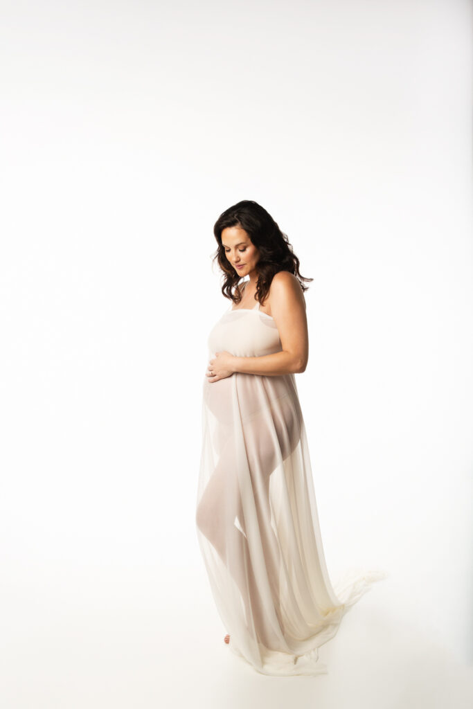 Caucasian mom to be, with dark wavy hair, is draped in ivory fabric sheer enough to show off her body shape for her pregnancy portraits, created in Fort Mill SC.