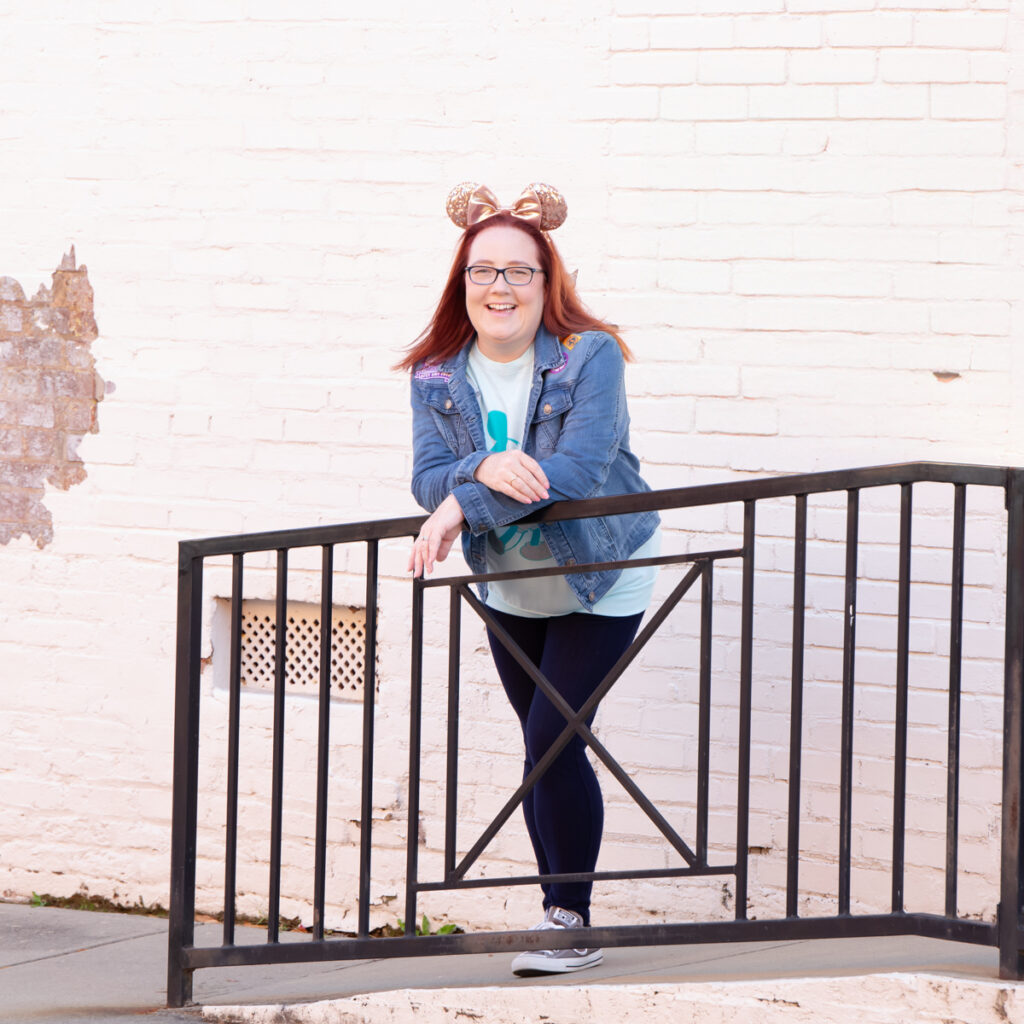 Disney planner posing outside off Main Street, Fort Mill SC for branding professional portraits. Woman is leaning over black fencing casually dressed smiling at the camera. 